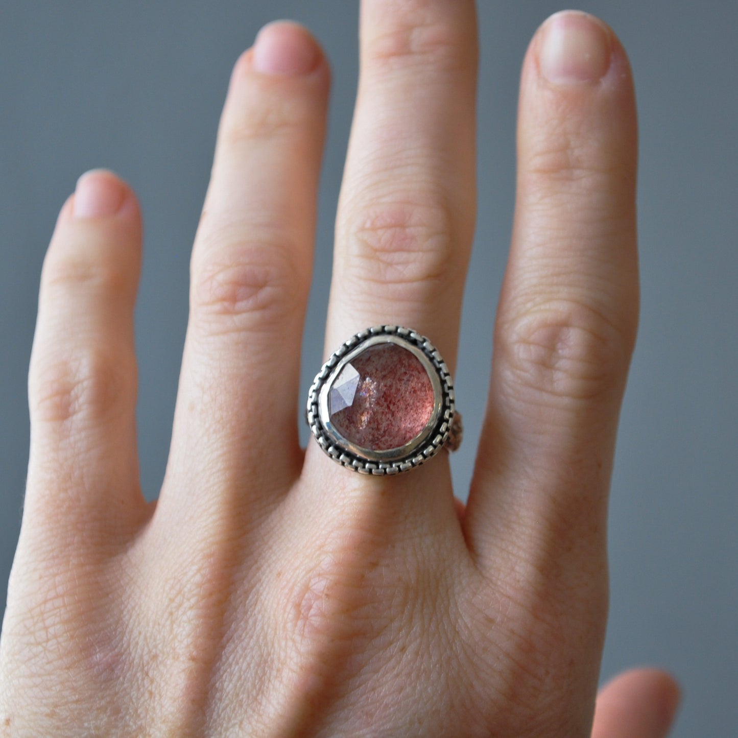 A silver ring with a round, faceted pinkish-red gemstone, worn on a person's ring finger against a neutral gray background.