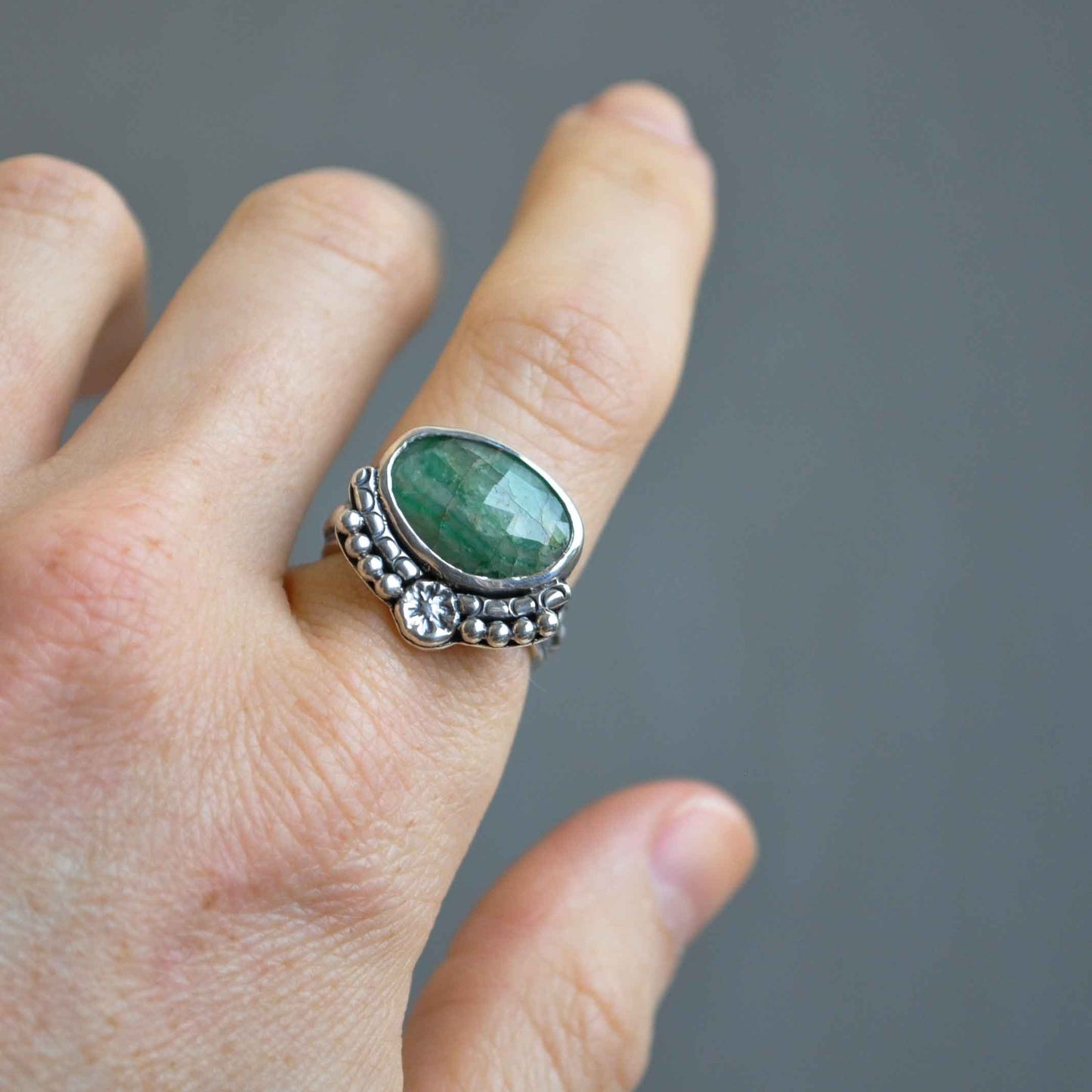 Hand wearing a silver ring with a green gemstone on a gray background