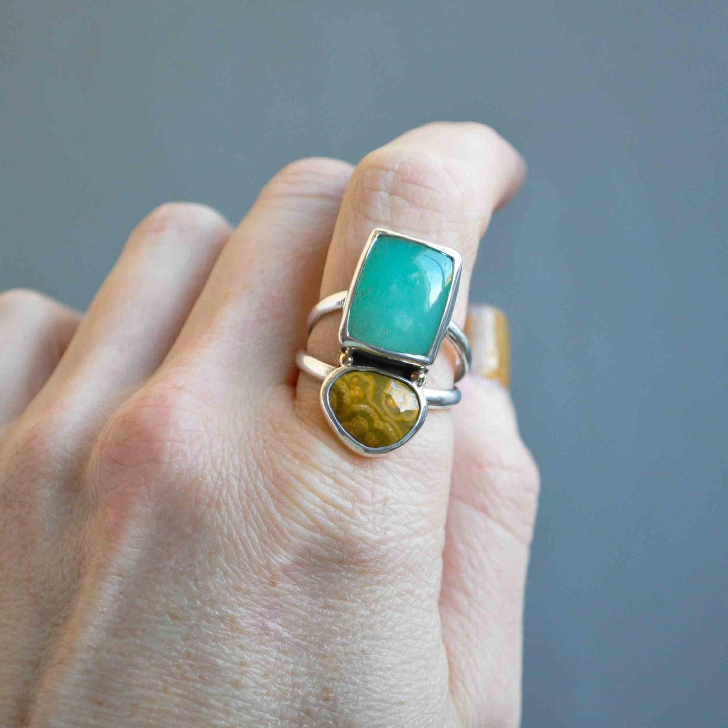 Hand wearing a ring with green and yellow gemstones on a gray background