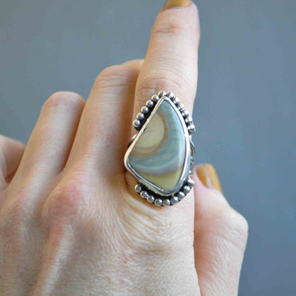 Hand wearing a large, decorative ring with a teardrop-shaped stone against a neutral background