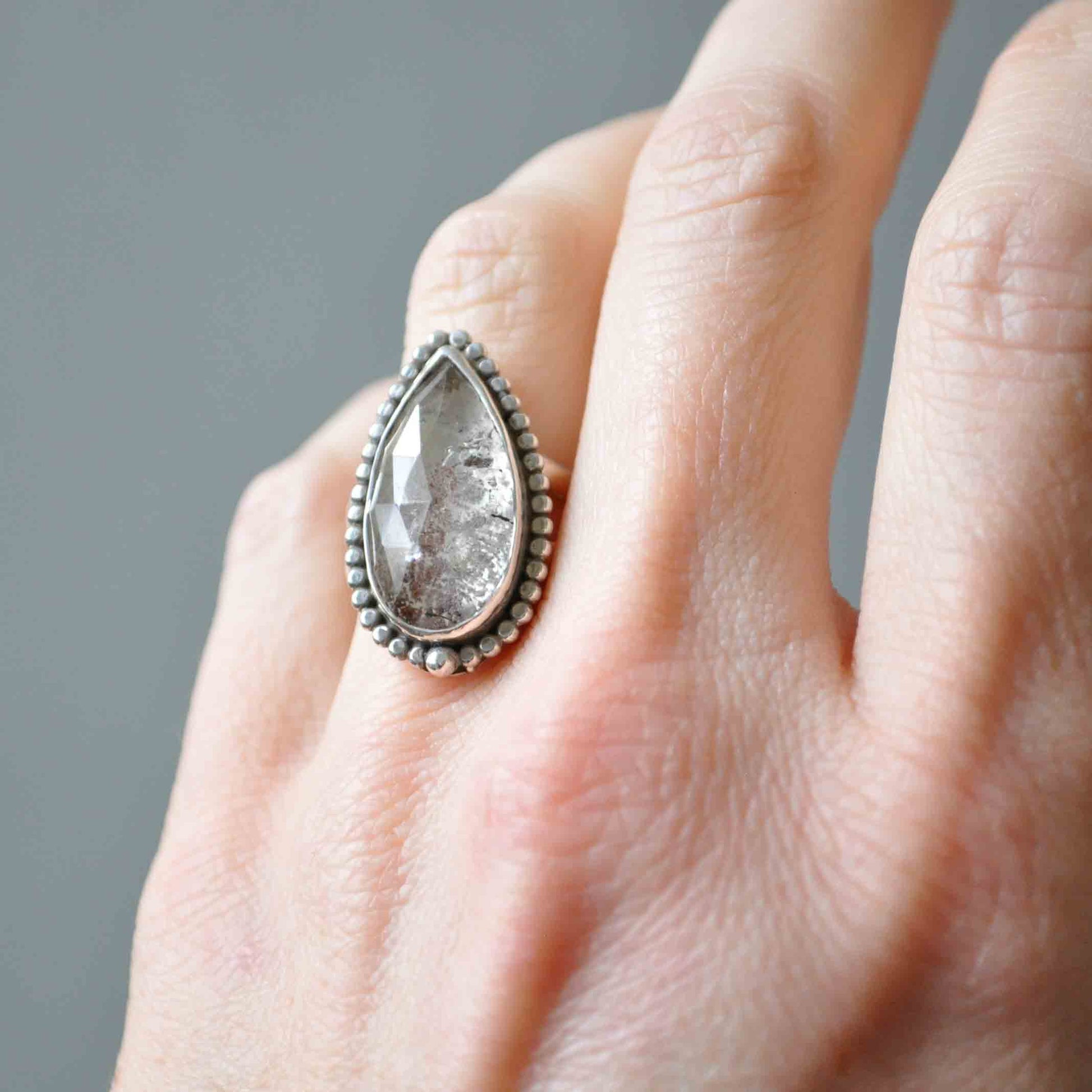 Teardrop-shaped silver ring on a finger against a gray background