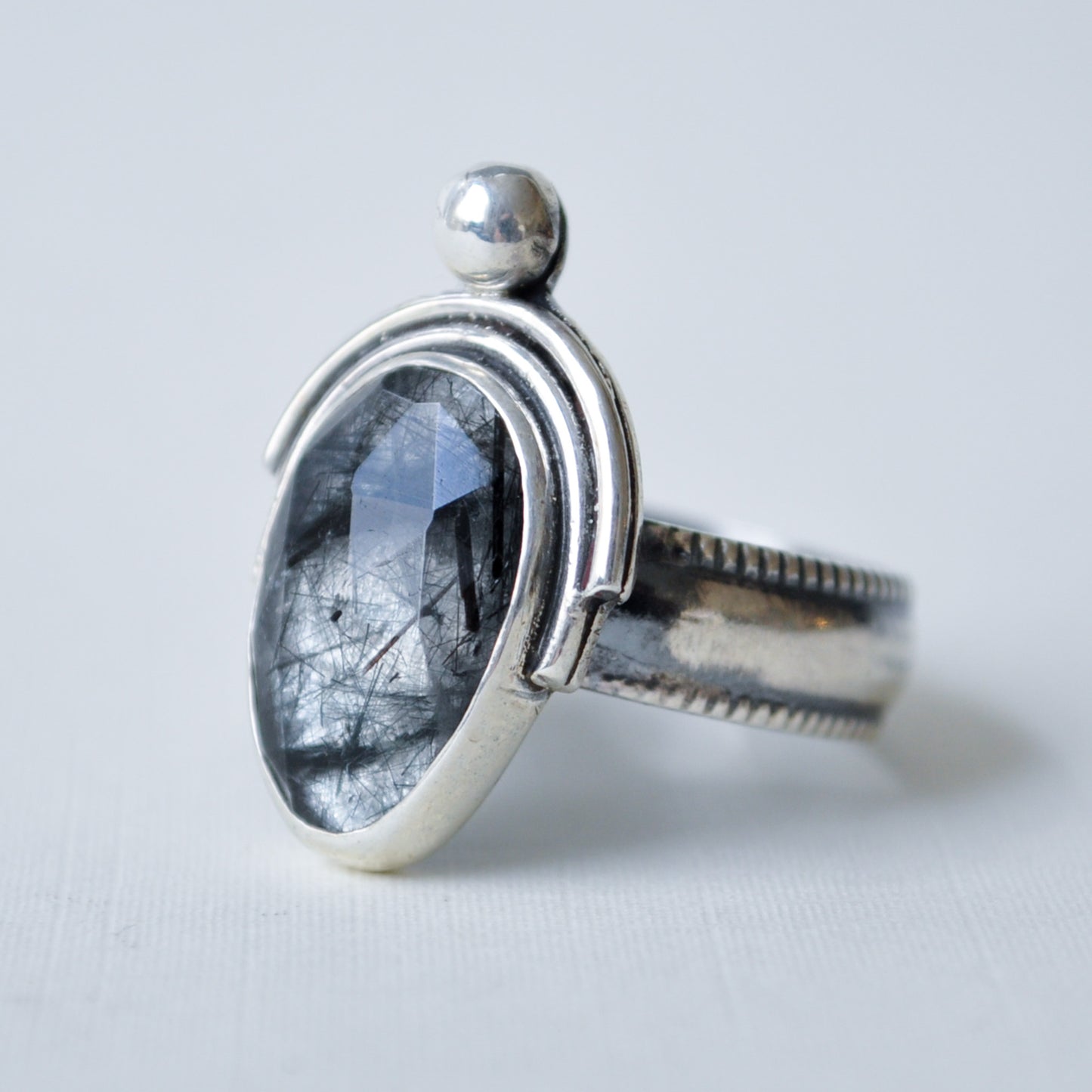 A sterling silver ring featuring an oval-cut black rutilated gemstone with visible hair-like inclusions, set in a polished silver bezel with a small spherical accent on top, photographed on a light gray background.