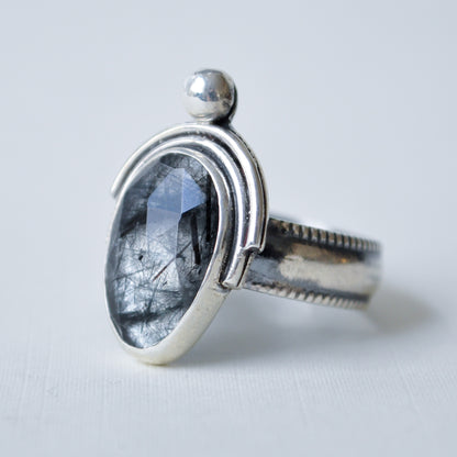 A sterling silver ring featuring an oval-cut black rutilated gemstone with visible hair-like inclusions, set in a polished silver bezel with a small spherical accent on top, photographed on a light gray background.