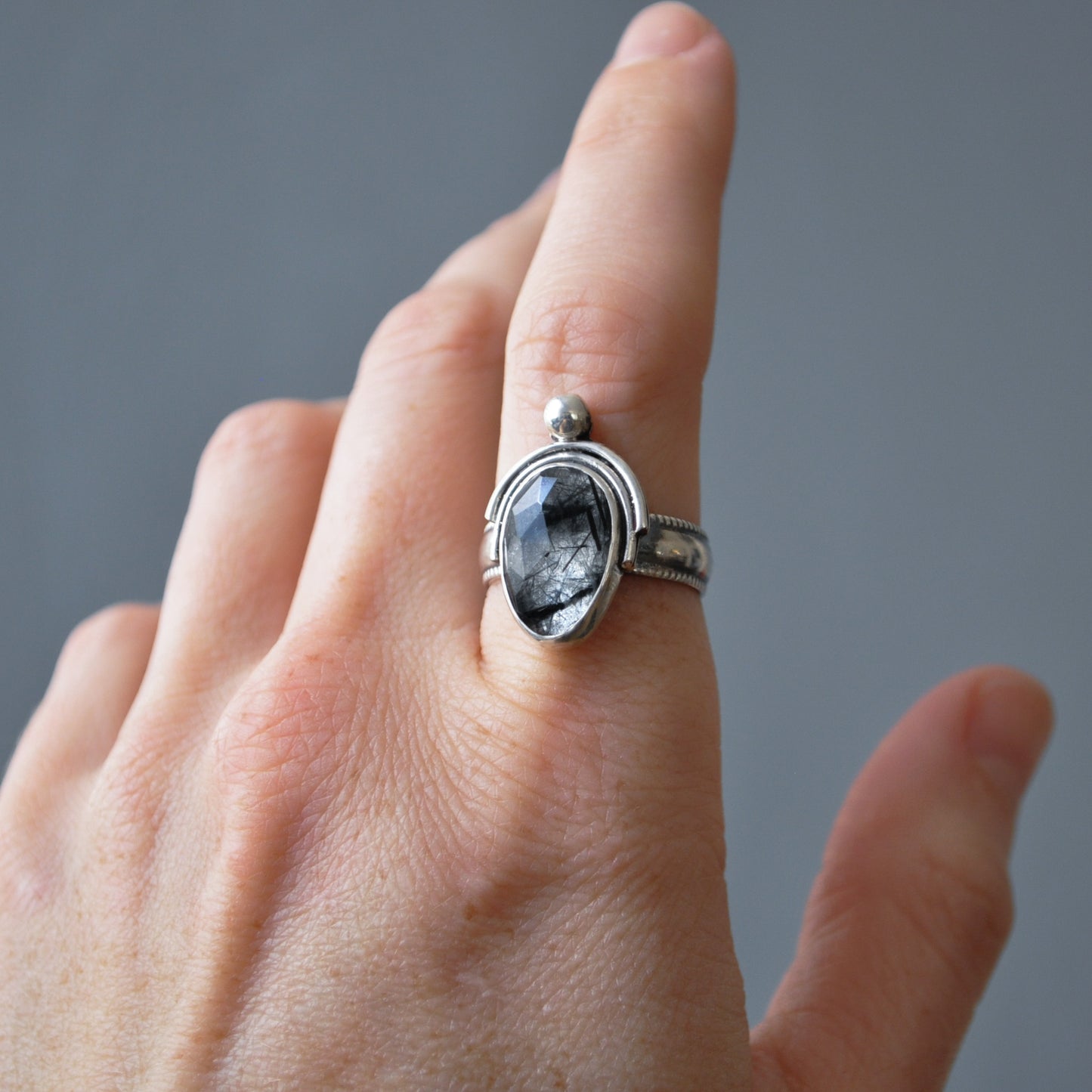 A silver ring with an oval dark gray stone featuring black inclusions, worn on a person's index finger against a neutral gray background.