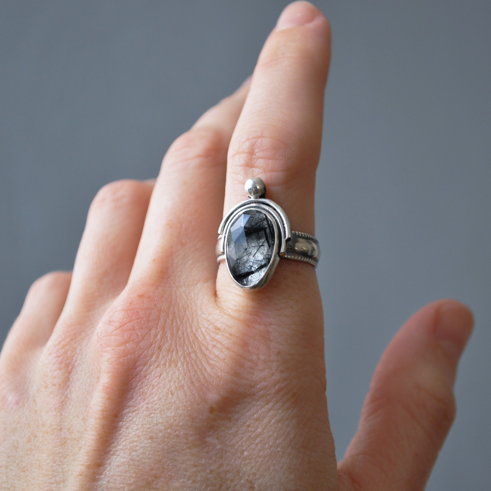 A silver ring with an oval dark gray stone featuring black inclusions, worn on a person's index finger against a neutral gray background.