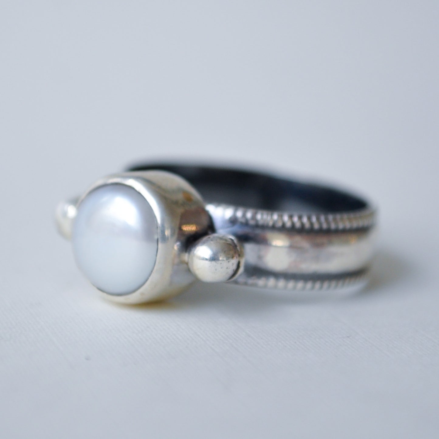 A sterling silver ring featuring a large, round, lustrous white pearl bezel-set on the front, with a textured beaded band and small decorative silver beads on either side, photographed on a light gray background.