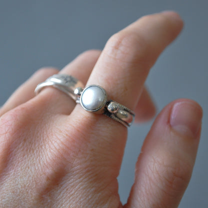 A close-up of a hand wearing a silver ring with a round white pearl-like center stone and a small clear stone accent, against a soft gray background.