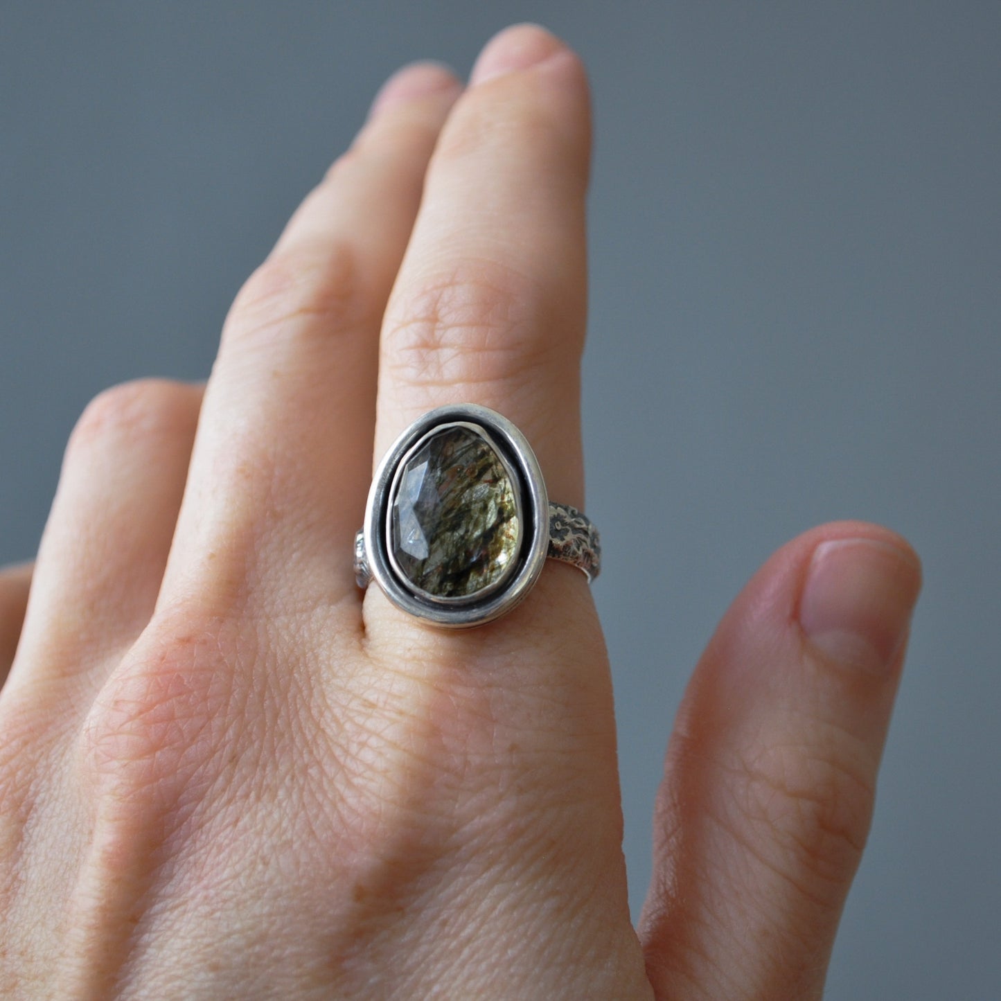 A silver ring with a large, oval, textured brown and gray stone set on a hand, shown against a solid gray background.