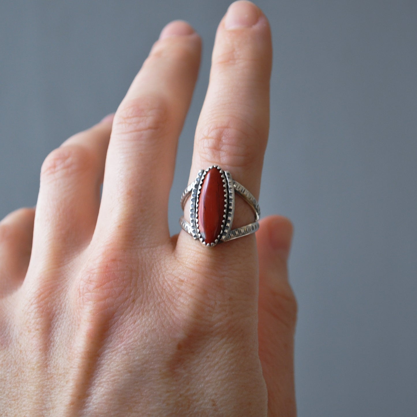 A silver ring with an oval red cabochon gemstone, worn on a person's finger against a neutral gray background.