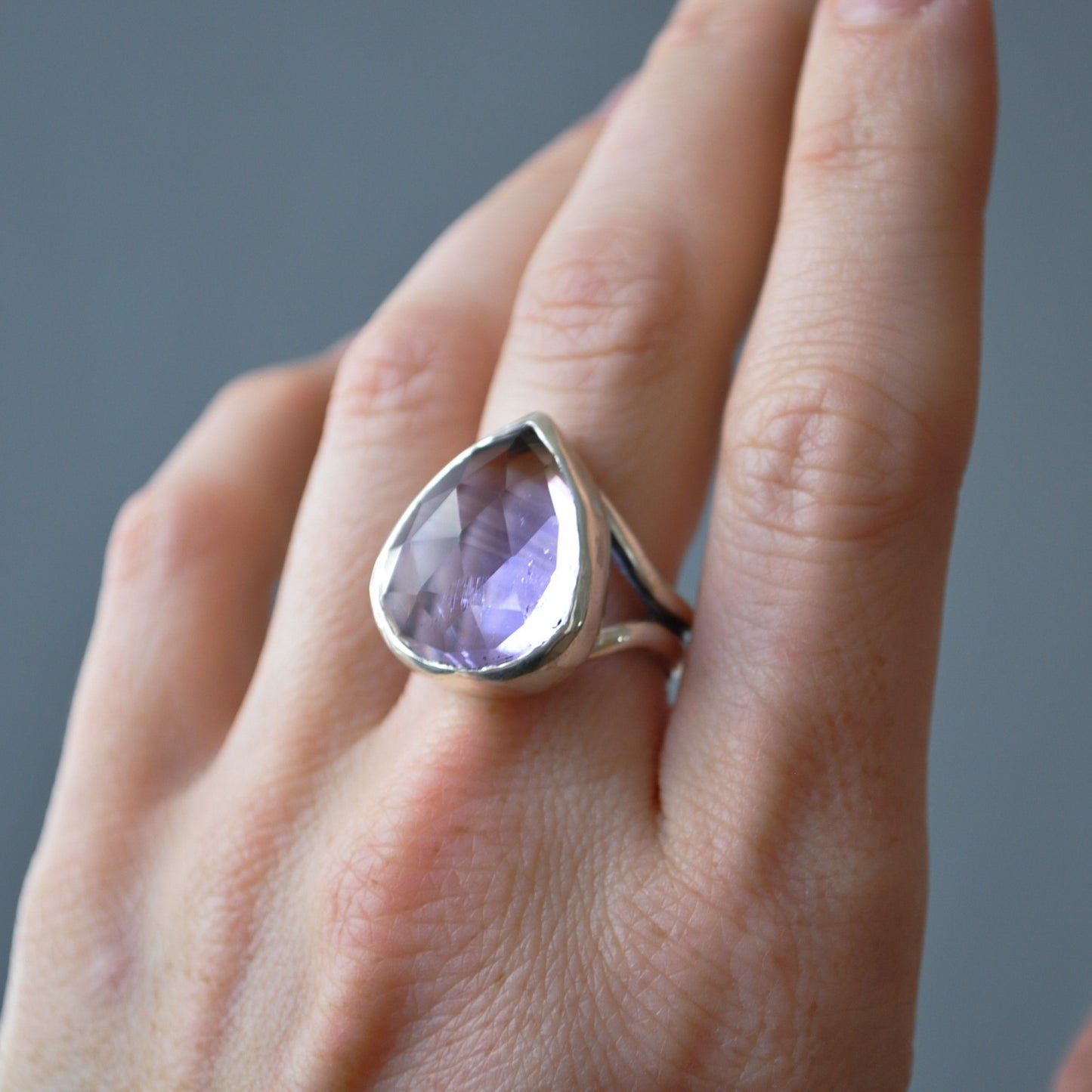 A silver ring featuring a large, teardrop-shaped faceted light purple gemstone, shown worn on a person's finger against a neutral gray background.