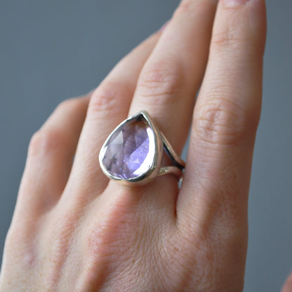 A silver ring featuring a large, teardrop-shaped faceted light purple gemstone, shown worn on a person's finger against a neutral gray background.