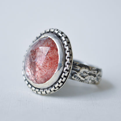 A silver ring featuring a large, oval, faceted pinkish-red gemstone with visible internal crystalline structure, set in an ornate, textured bezel and band.
