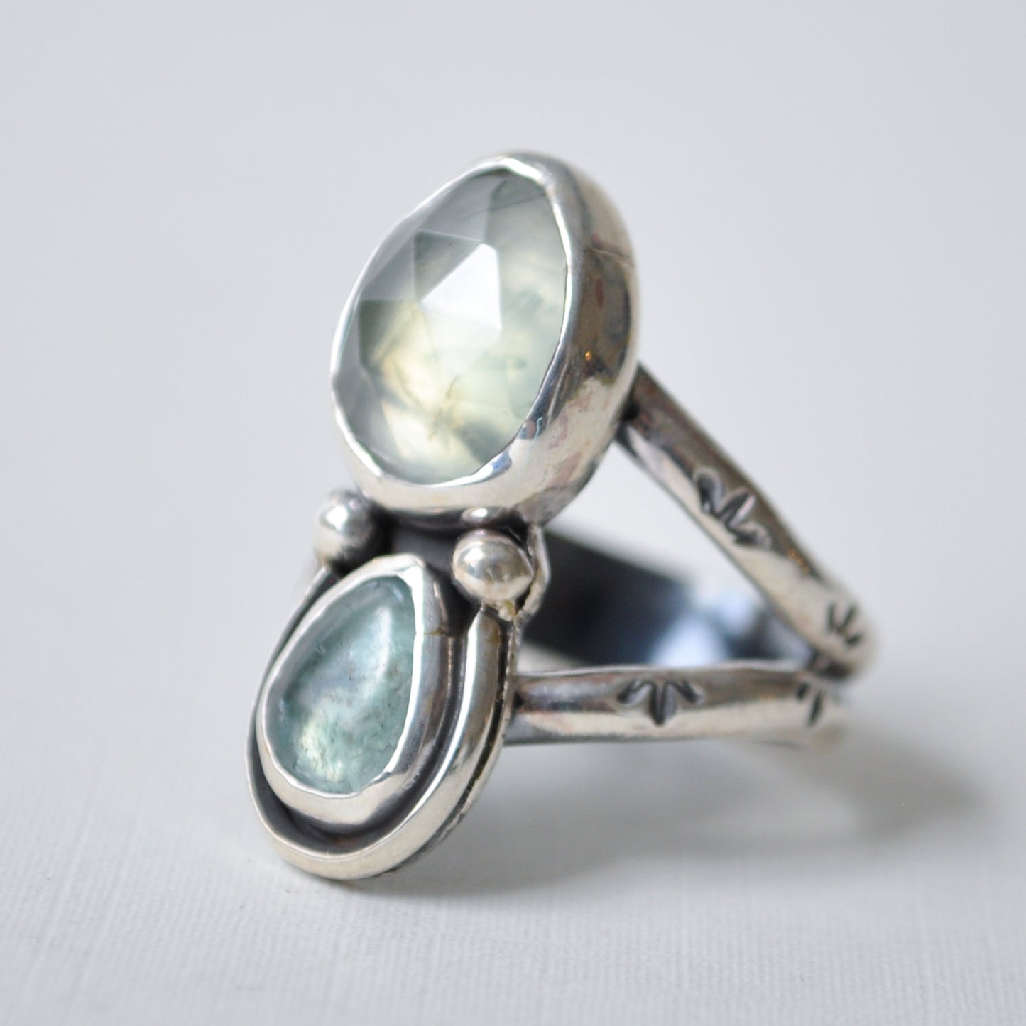 A sterling silver ring featuring two teardrop-shaped light blue-green gemstones, one faceted oval stone and one teardrop stone, set against a light gray background.