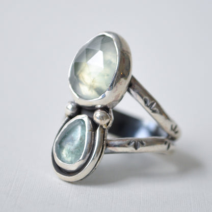 A sterling silver ring featuring two teardrop-shaped light blue-green gemstones, one faceted oval stone and one teardrop stone, set against a light gray background.
