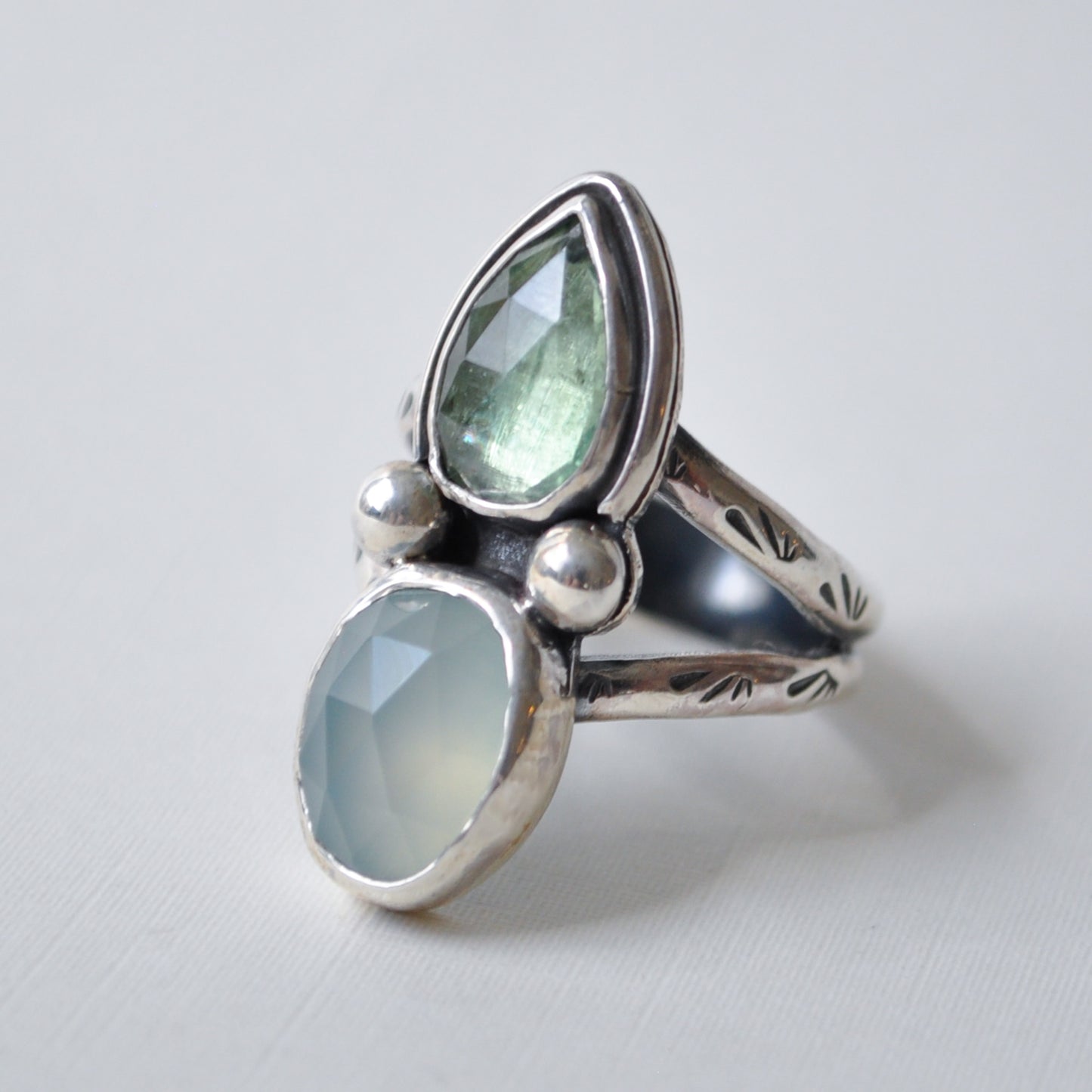 A sterling silver ring featuring two faceted gemstones, a larger teardrop-shaped light green stone and a smaller oval-shaped pale blue-green stone, displayed on a light gray background.