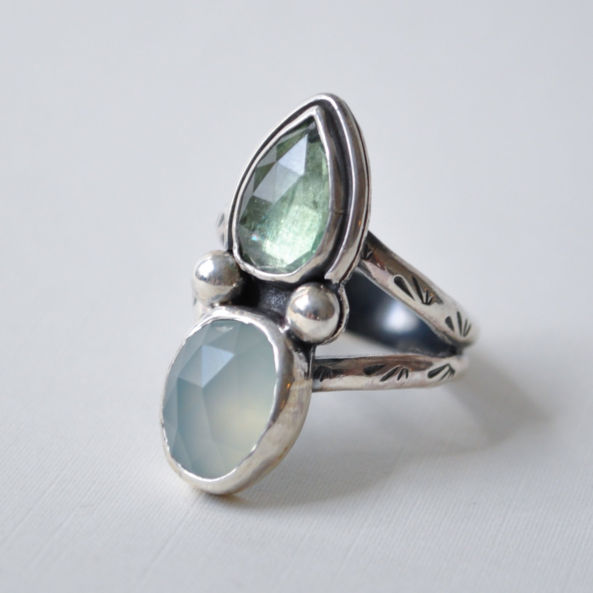 A sterling silver ring featuring two faceted gemstones, a larger teardrop-shaped light green stone and a smaller oval-shaped pale blue-green stone, displayed on a light gray background.