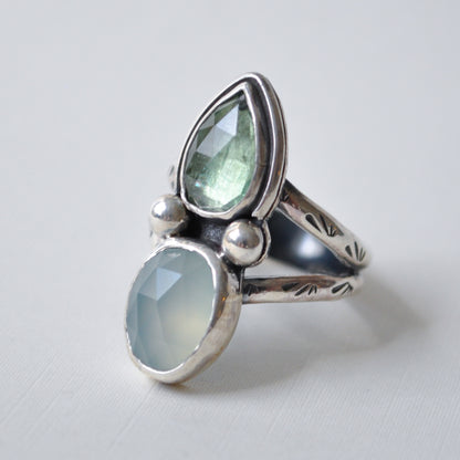 A sterling silver ring featuring two faceted gemstones, a larger teardrop-shaped light green stone and a smaller oval-shaped pale blue-green stone, displayed on a light gray background.