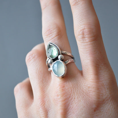 A close-up of a hand wearing a silver ring with two light green stones, one teardrop-shaped and one oval, set against a soft gray background.