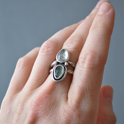 A close-up of a hand wearing a silver ring with two oval green stones set in a stacked design, against a soft gray background.