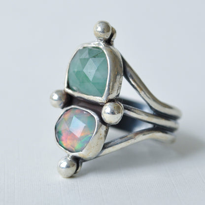 A sterling silver ring featuring two faceted gemstones, one light green and one iridescent opal with rainbow reflections, set in a split-band design on a light gray background.