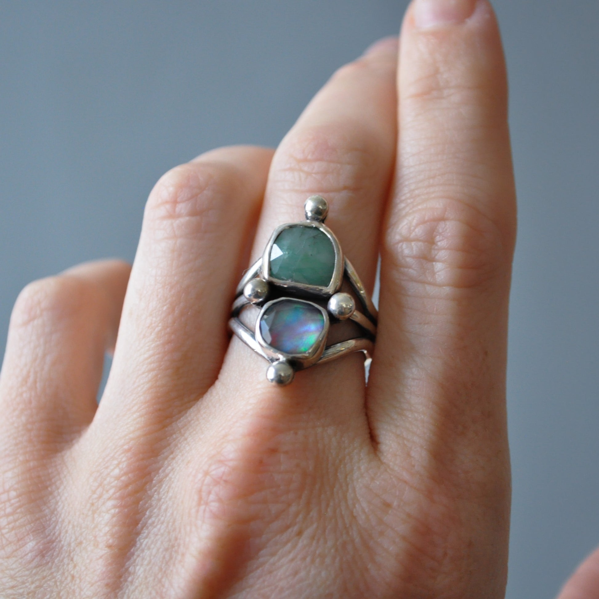 A sterling silver ring with a green stone and an iridescent opal, worn on a person's finger against a neutral gray background.