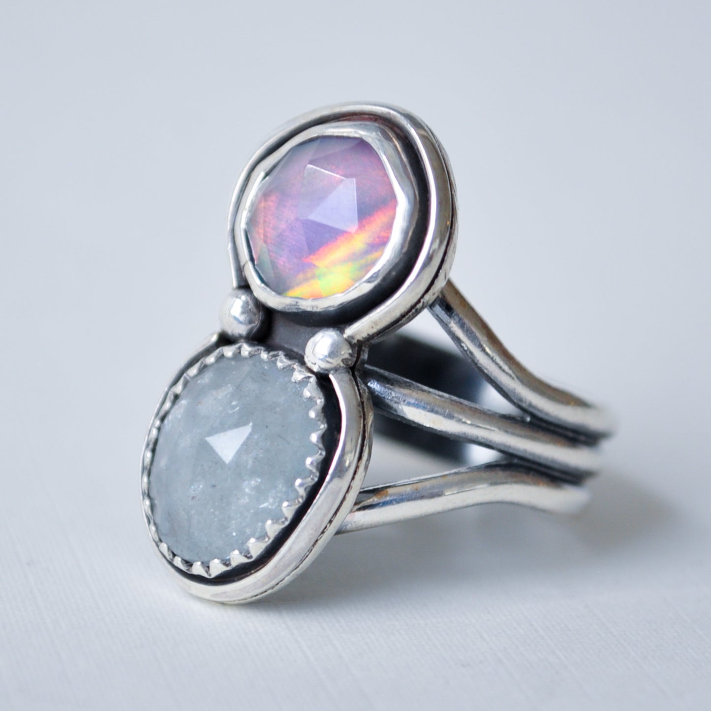 A sterling silver ring featuring two oval gemstones, one with a vibrant iridescent rainbow sheen and the other a smooth light gray stone, set against a clean light gray background.