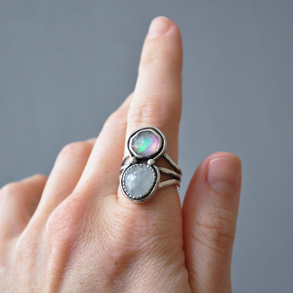 A close-up of a hand wearing a silver ring with two oval stones, one iridescent and one milky white, against a soft gray background.