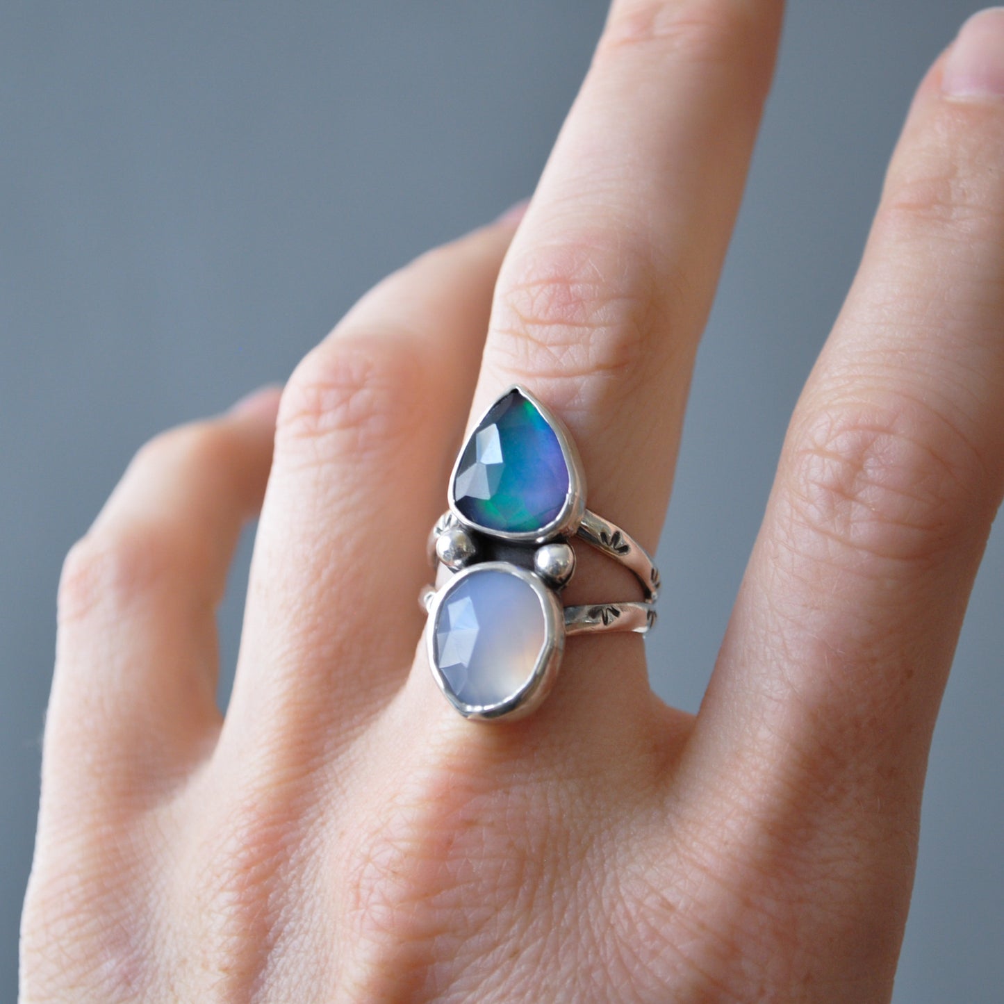 A sterling silver ring with two stones worn on a finger. The top stone is a teardrop-shaped iridescent blue-green opal, and the bottom stone is a round milky white moonstone, set on a silver band with small decorative beads.
