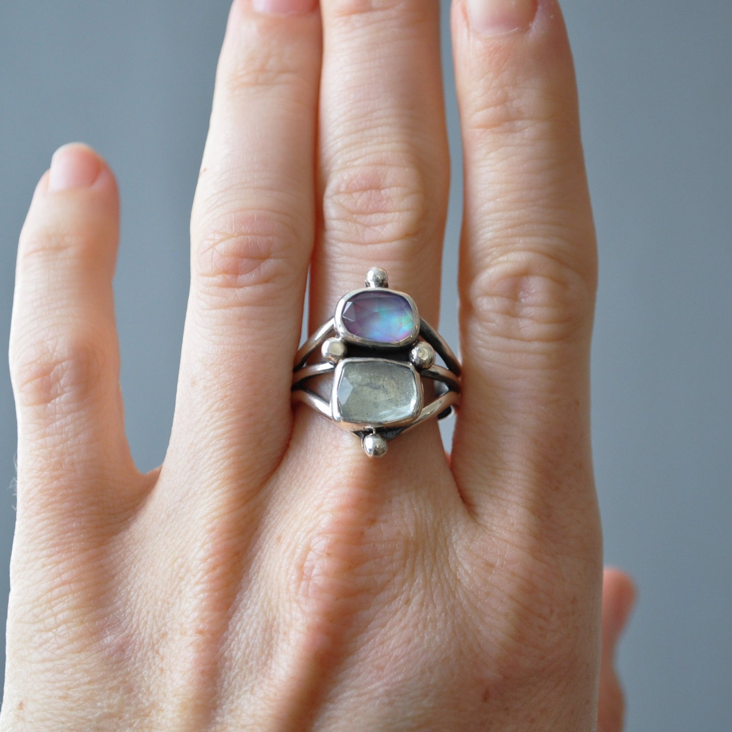A sterling silver ring with two iridescent stones, one blue and one green, worn on a hand against a soft gray background.