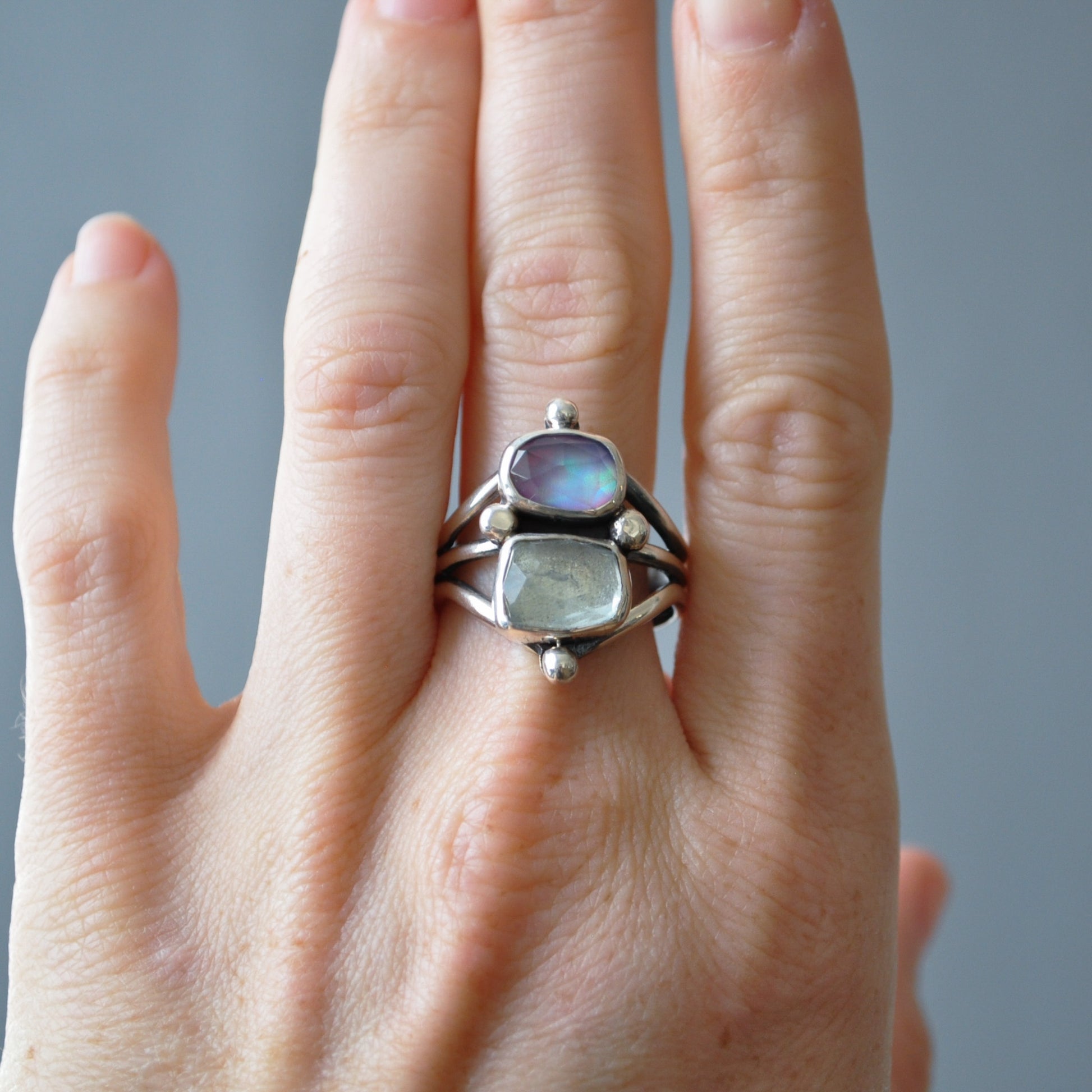A sterling silver ring with two iridescent stones, one blue and one green, worn on a hand against a soft gray background.